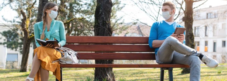 Two people with masks sit far apart from each other on a bench to help prevent the spread of COVID.